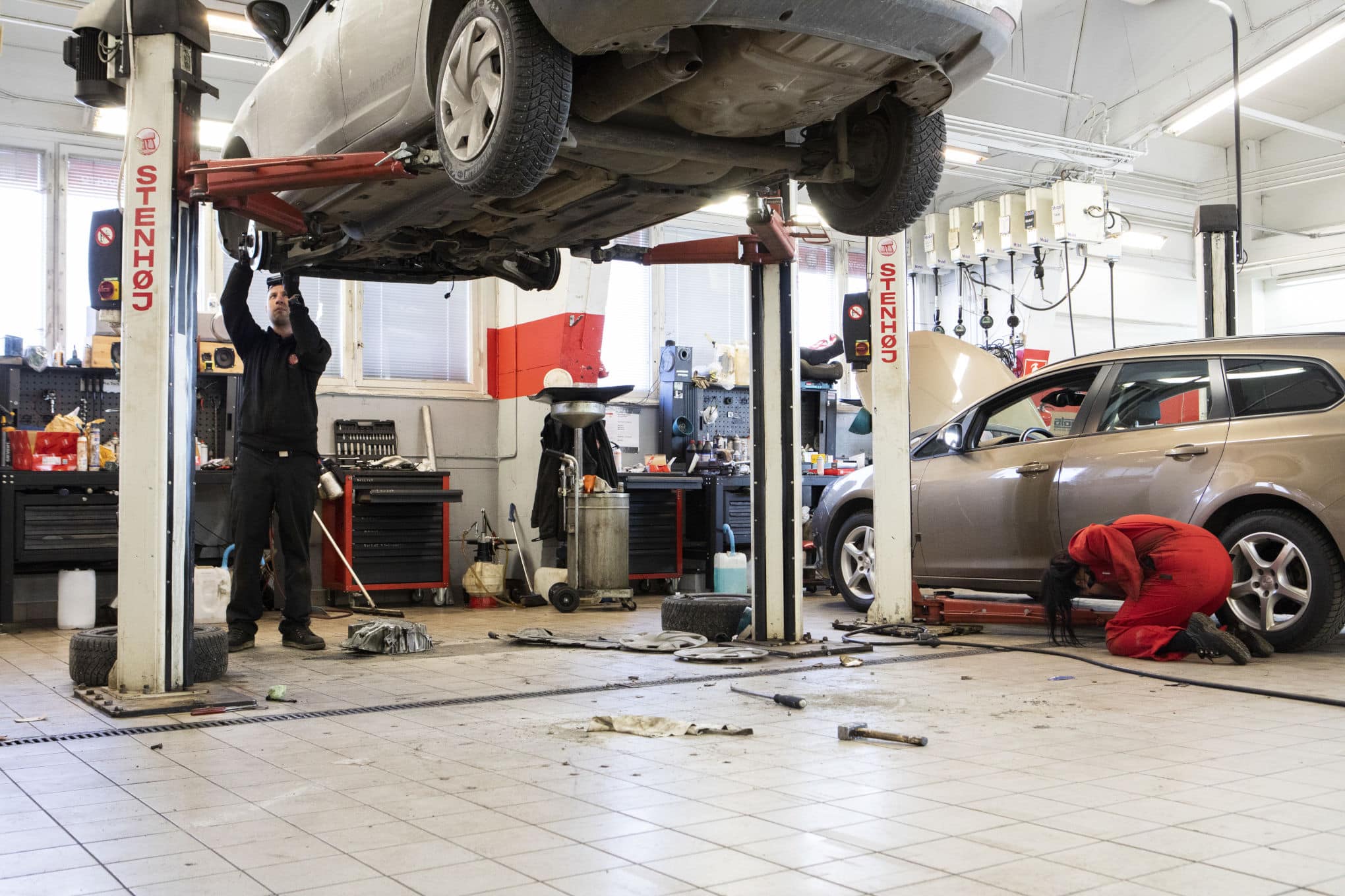 Autokorjaamossa on auto, joka on nostettu ilmaan. Auton vieressä on työntekijä ja muita autoja.