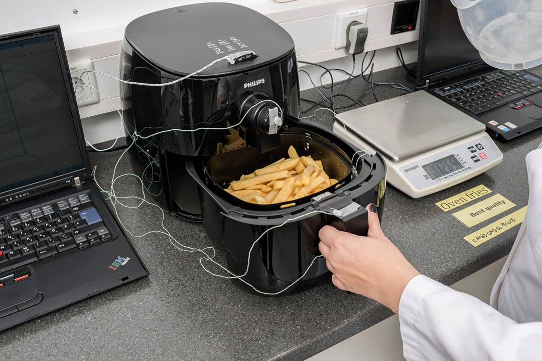 Henkilö avaamassa airfryeriä laboratoriossa. Airfryerin sisällä on ranskalaisia perunoita ja sen ulkopintoihin on kiinnitetty lämpömittareita.