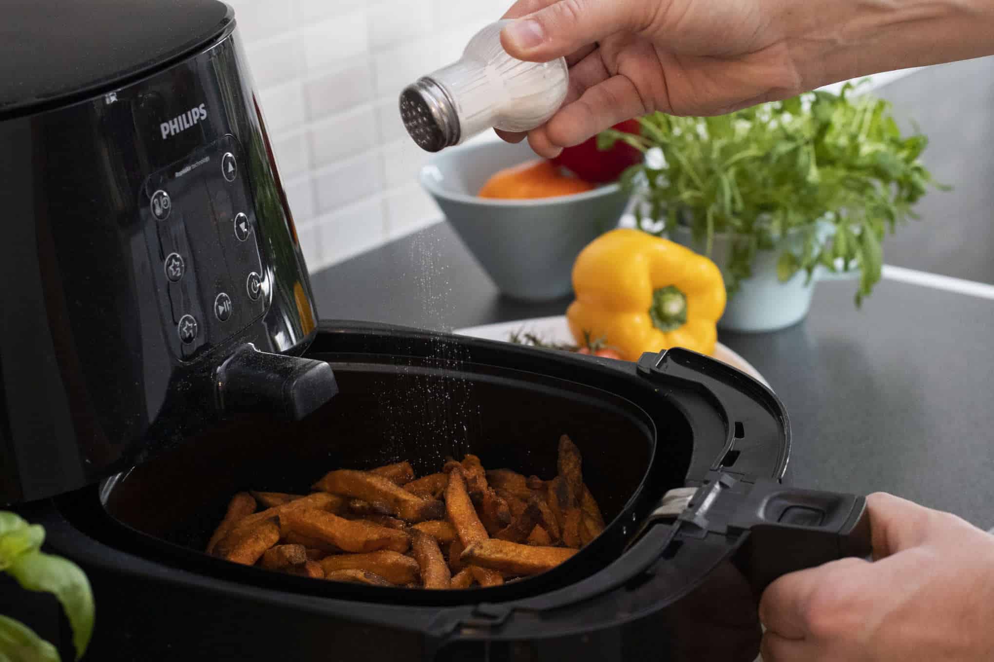 Henkilö sirottelee suolaa airfryerissä olevien bataattiranskalaisten päälle.