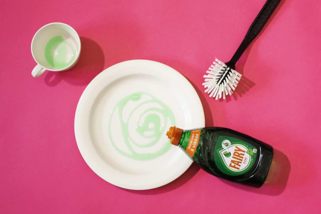 Kahvikuppi, lautanen, tiskiharja ja Fairy-merkkinen tiskiainepullo.