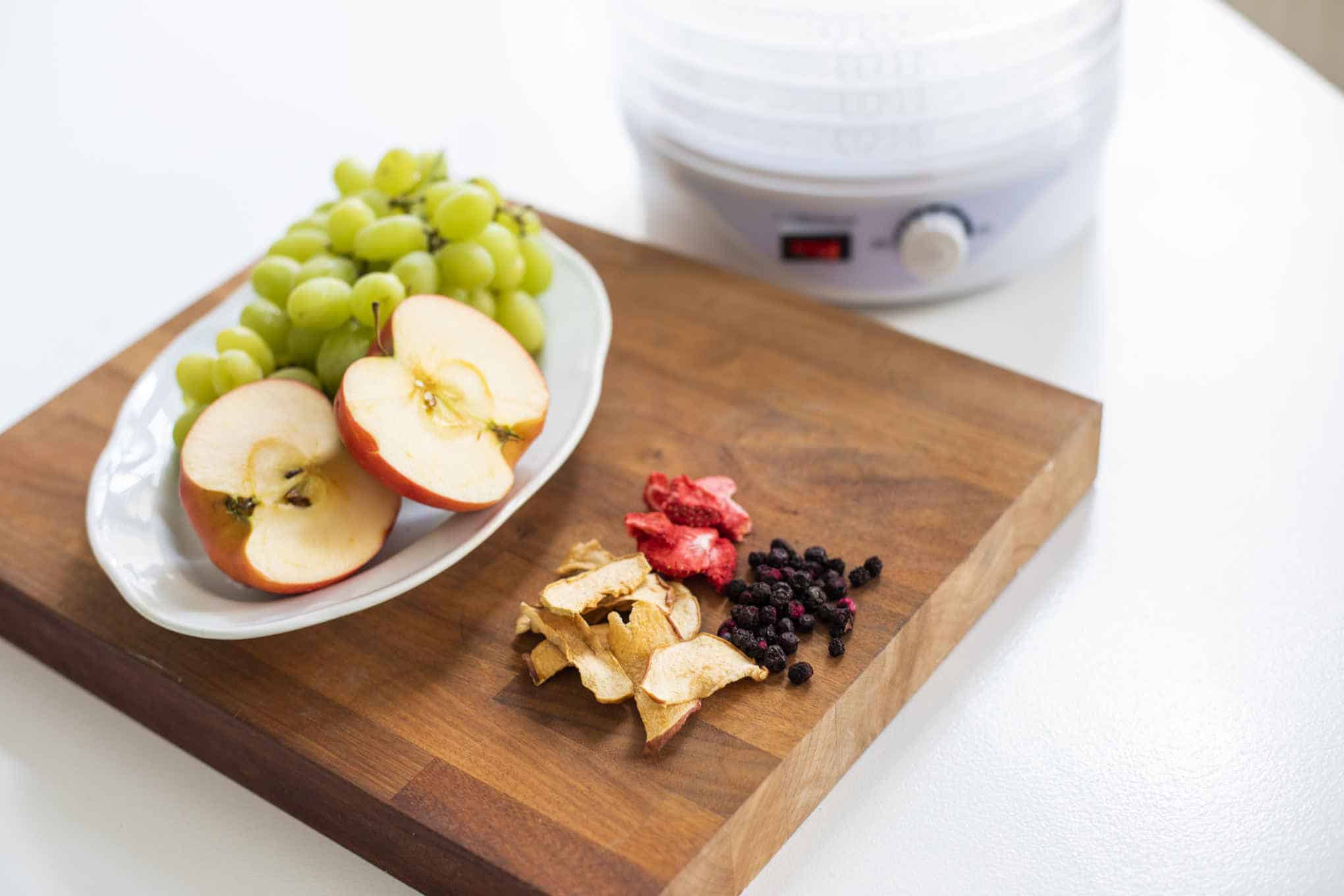 Kasvikuivuri ja leikkuulauta, jonka päällä on tuoreita ja kuivattuja hedelmiä.