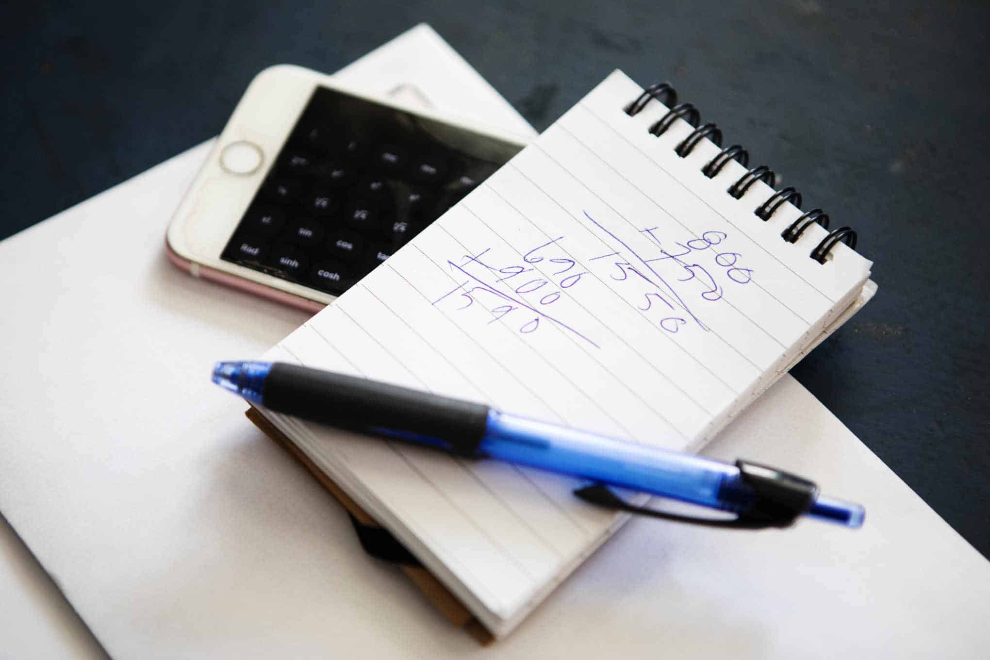 Puhelin, kynä ja muistilehtiö, jossa on laskelmia.