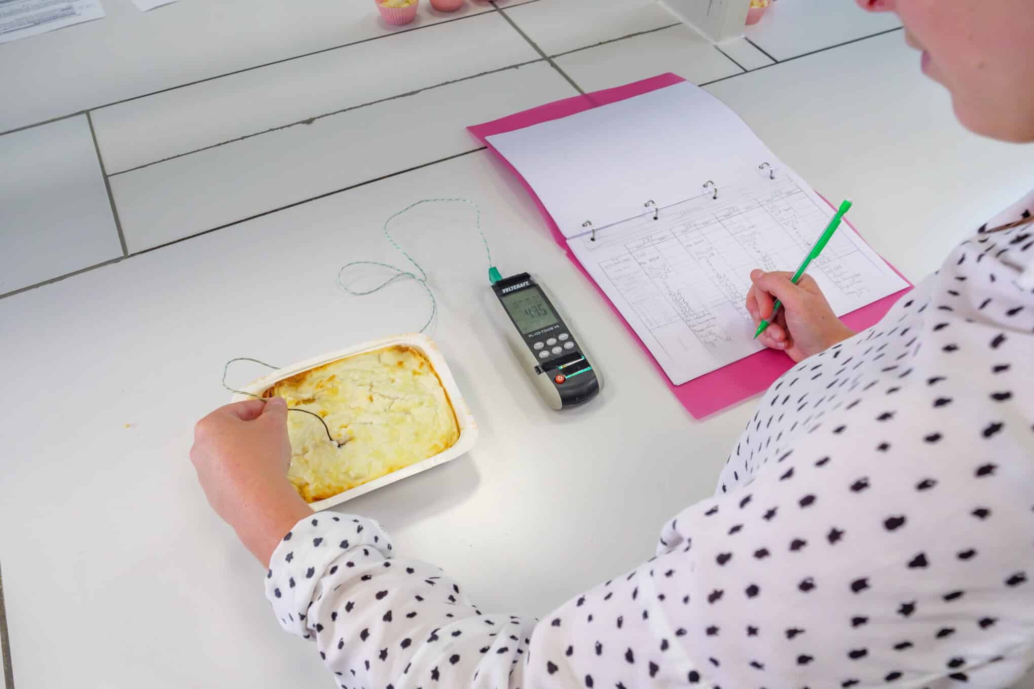 Henkilö mittaamassa lasagnen lämpötilaa lämpömittarilla ja merkitsemässä tuloksia monisteeseen.