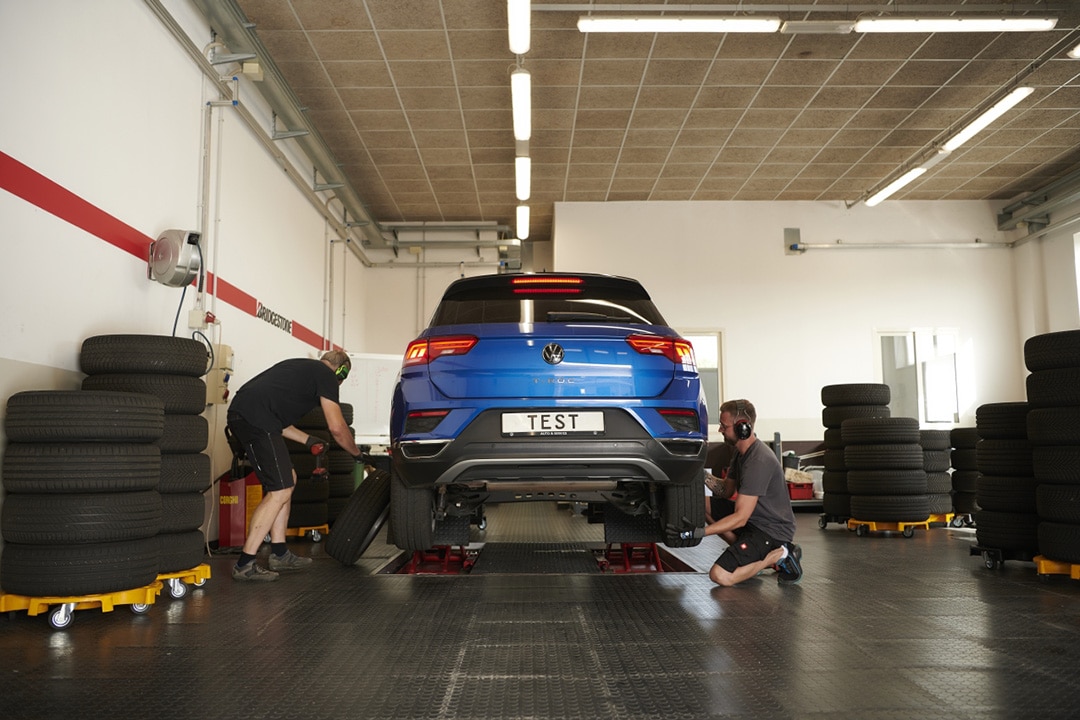 Kaksi henkilöä vaihtaa auton renkaita autotallissa. Auton vieressä on rengaspinoja.