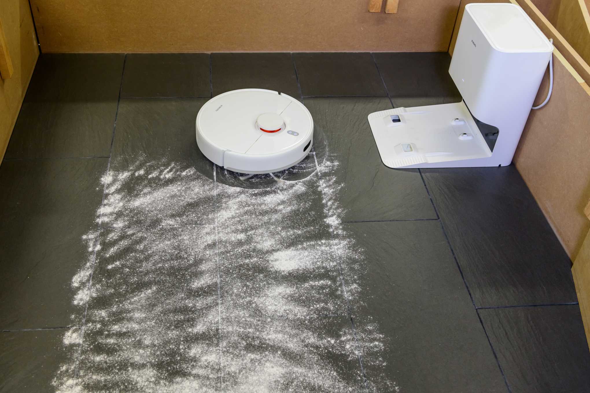 Huoneen lattialla on robotti-imuri ja dolomiittihiekkapölyä. Seinään on kiinnitetty robotti-imurin latausasema.