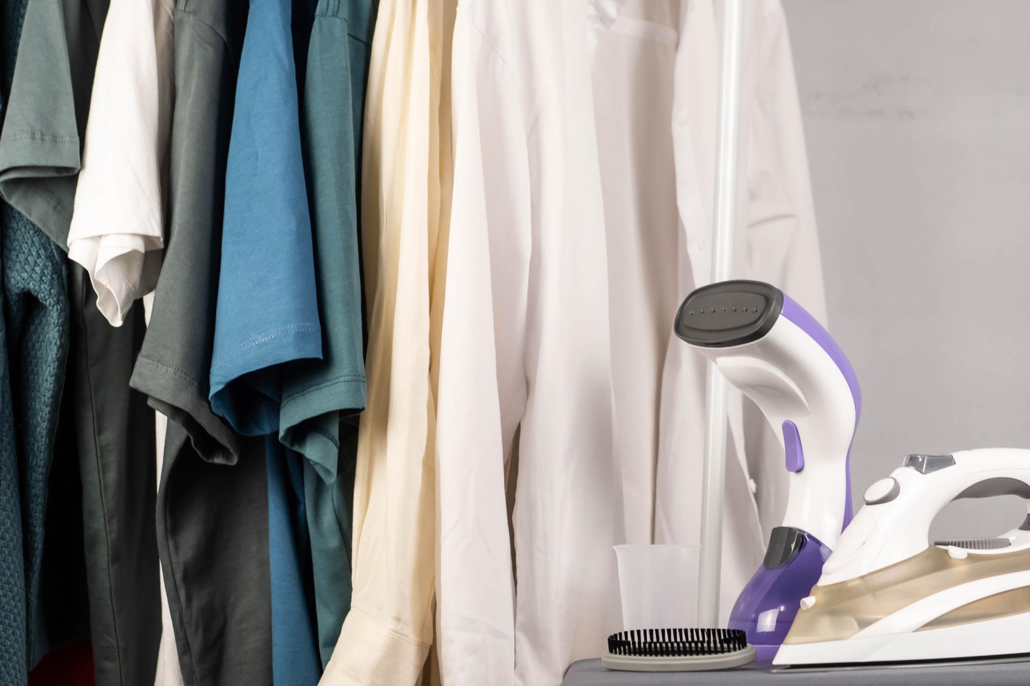 Vaatehöyrystin, silitysrauta ja henkarissa roikkuvia vaatteita.